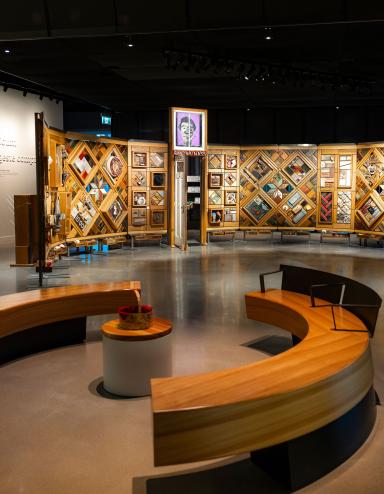 A museum exhibition featuring a large, curved freestanding wooden wall on which is mounted numerous small objects arranged in diamond and rectangular patterns. In the foreground, there circular benches creating an inviting space for visitors to view and contemplate the exhibit. Partially obscured.