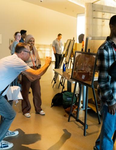 Des projets d’élèves sont exposés sur des chevalets dans une galerie de musée. Deux élèves parlent de leur travail avec des adultes. Partially obscured.