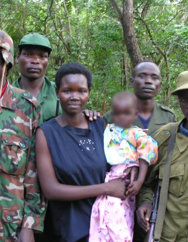 A woman is holding a baby, while four men in army fatigues stand beside and behind her. They are all standing in front of a forest, posed for the camera. Partially obscured.