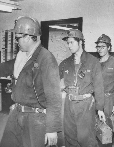 A line of miners hold lunch pails as they punch a time clock. Partially obscured.