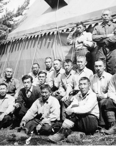 A group of men pose for a picture with a large tent and a tree in the background. Partially obscured.