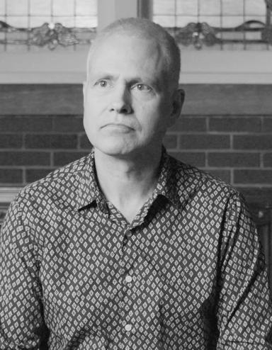 Delwin Vriend sits in a chair with a neutral expression on his face. He is lit from the front as if he is doing an interview on camera. Partially obscured.