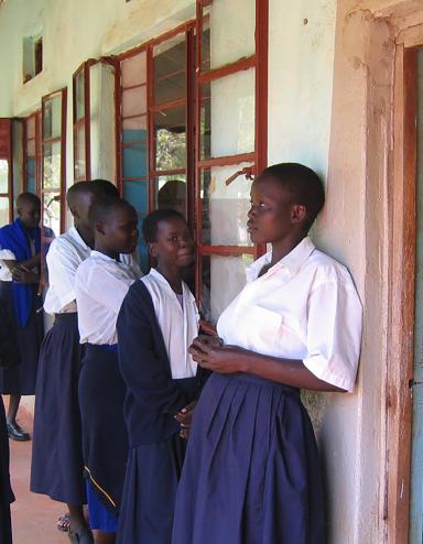 A group of young women and girls in their teens stand together on the front porch of a building. Most are dressed in white blouses and blue skirts, with two girls also wearing blue sweaters. To the right, a doorway reveals a roomful of students sitting at wooden desks and writing on notepads. Partially obscured.