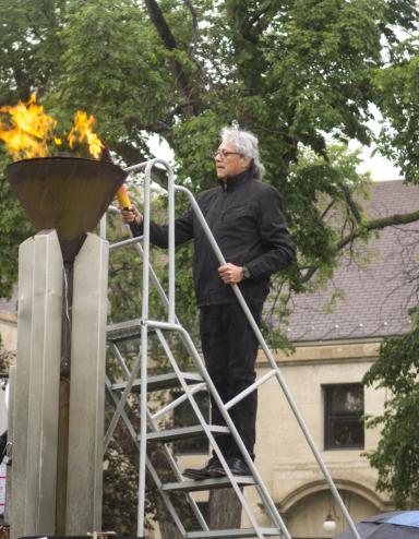 A man carrying a torch stands on a ladder to light a flame within a copper cauldron. Partially obscured.