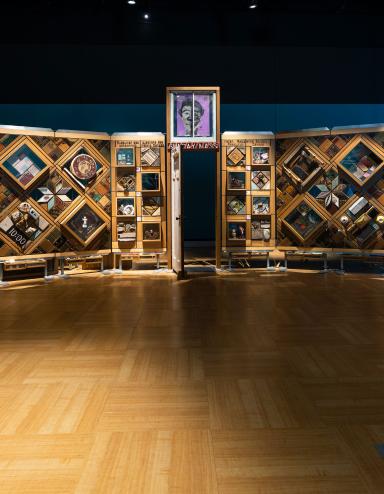 A large, curved freestanding wooden wall on which is mounted numerous small objects arranged in diamond and rectangular patterns. There is an open door in the centre of the wall. Partially obscured.