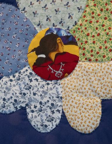 Close-up of a flower on a quilt. Each of the petals is a different flowery fabric, while at the centre of the flower is fabric onto which is printed the face of an Indigenous woman in profile. Partially obscured.