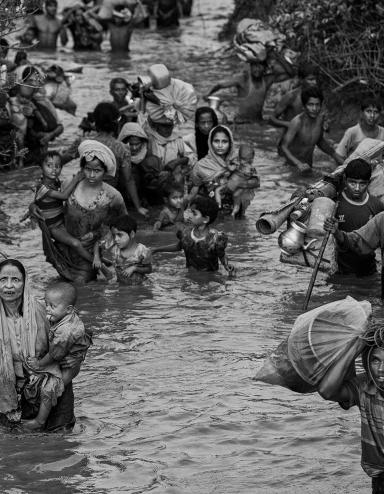 Rohingya women, children and men wade through waist-high muddy river water. Some carry young children, while others carry bags of possessions, including household items. Partially obscured.