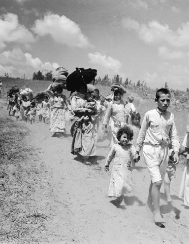 Women and children carrying bundles on their heads walk along a dirt road. Partially obscured.
