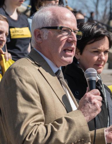 A middle-aged man with short grey hair stands with a mic speaking to a crowd. Partially obscured.