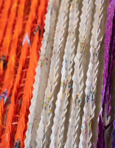 Multi-coloured origami cranes strung together vertically and hung on a wall. Partially obscured.