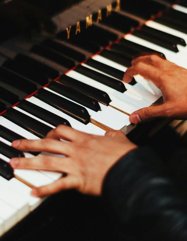 Two hands play the keys of a gleaming black Yamaha piano. The pianist, barely shown, wears a long-sleeved black top. Partially obscured.