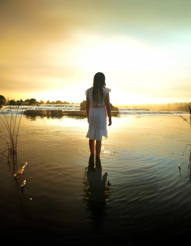 A girl with long dark hair wearing a knee-length white dress faces away, standing ankle deep in a calm lake, with the setting sun casting a warm yellow glow across the water. Partially obscured.