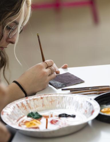 A young woman sitting at a table paints a design on a tile while a tin tray with various paints sits in front of her. An open bottle of red paint sits on the table. Partially obscured.
