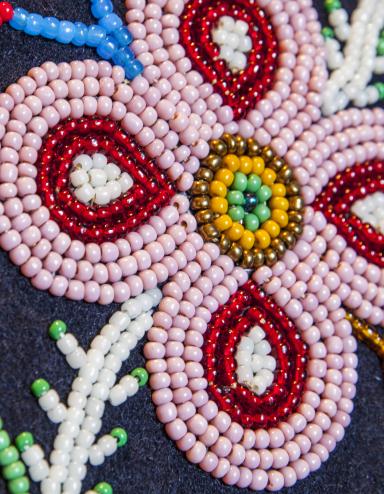 Close-up of a beaded floral design on dark fabric, featuring a pink four petal flower with red and white details, a yellow and green beaded center, and surrounding beadwork resembling leaves and stems. Partially obscured.