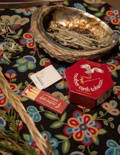 Smudging items arranged on a round table covered with a floral cloth, including an abalone shell holding dried sage, a feather, braided sweetgrass, cedar branches, and a small box of matches. Partially obscured.