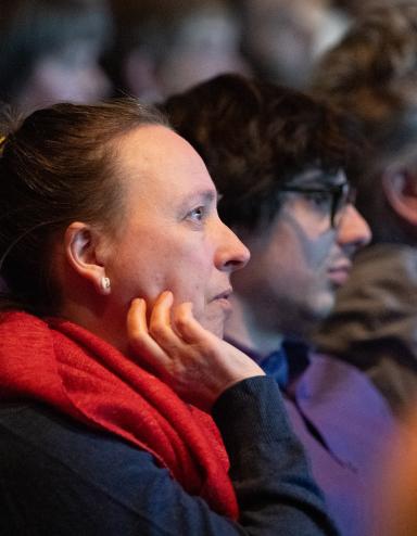 Audience members sit in a dimly lit venue watching a presentation, their faces illuminated by stage light. In the foreground, a person with their hair in a bun and wearing a bright red scarf rests their chin on their hand, looking attentively toward the stage. Other attendees sit beside them in soft focus, creating a quiet, contemplative atmosphere as the crowd listens. Partially obscured.