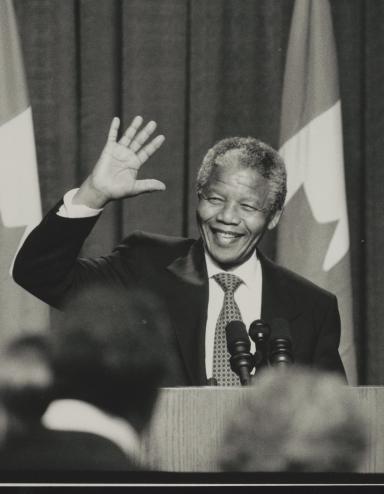 A black and white photograph of Nelson Mandela standing in front of a podium. He is smiling and waving. Partially obscured.