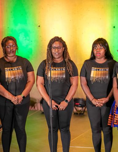 A group of six black performers standing on stage with colourful backlighting. Partially obscured.