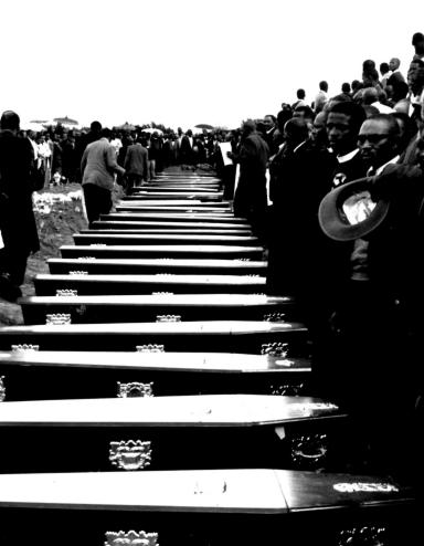 People stand in front of a row of coffins. Partially obscured.