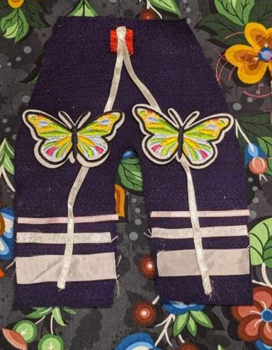 Pieces of crafted Indigenous- style clothing, including an orange shirt, pair of pants and made from colourful fabric and ribbon is are laid against a piece of craft paper adorned with flowers and berries. The clothing is includes an orange shirt, a pair of pants with ribbons and a pink tunic decorated with colourful fabric, appliqués and ribbons. The tunic has with a feathered shawl on the shoulders. Partially obscured.