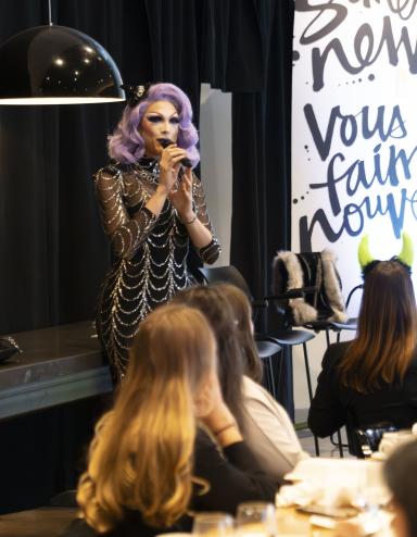 A drag performer with purple hair and a silver and black dress speaks in front of a group having brunch. Partially obscured.
