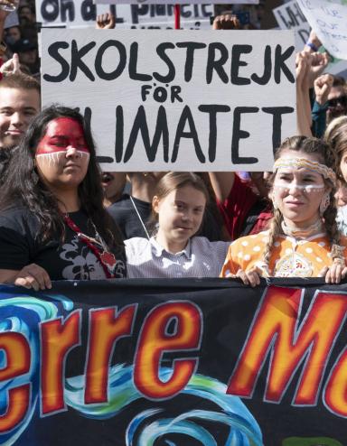 A crowd of youth hold protest signs and stand behind a large banner that reads “La Terre Mère,” or “mother earth” in English. Partially obscured.