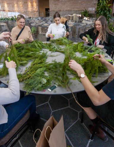 A group of people sit at a round table handling wreath materials. Partially obscured.