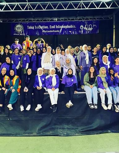 Dozens of people are gathered on a stage, most smiling and wearing the colour purple. Many are wearing hijabs. A banner hangs at the back of the stage that reads "Take Initiative. End Islamophobia. #OurLondonFamily." Partially obscured.