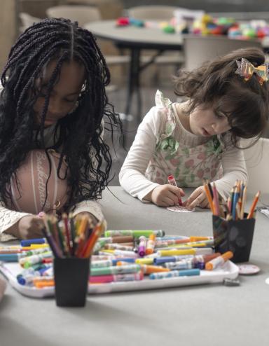 Two young children sit side by side at a round table, engaged in an art activity with markers and coloured pencils. Partially obscured.