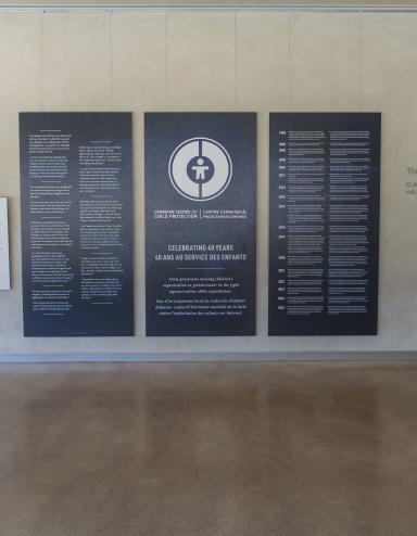 Large dark blue panels with white text and graphics displayed in the Community Corridor at the Canadian Museum for Human Rights. The centre panel features a circular logo and commemorates the 40th anniversary of the Canadian Centre for Child Protection. The two panels on either side present bilingual text in English and French. Partially obscured.
