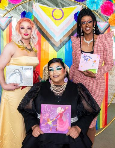 Three drag artists holding books are standing in front of a rainbow-themed circle arch. Partially obscured.