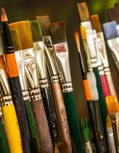 Various sizes of paint brushes standing in a paint-brush holder against a blurred green backdrop. Partially obscured.