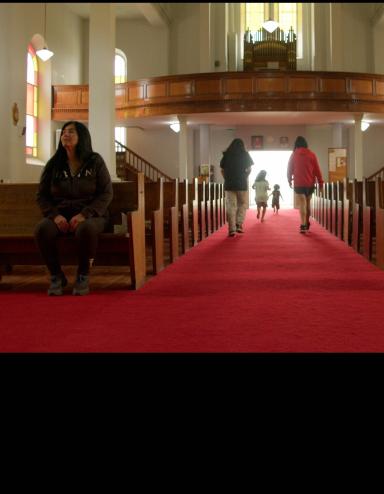 A church interior with a long red-carpeted aisle between wooden pews. Sunlight filters through stained-glass windows as several children run toward the bright entrance at the far end while an Indigenous woman sits alone in a pew to the left. Partially obscured.