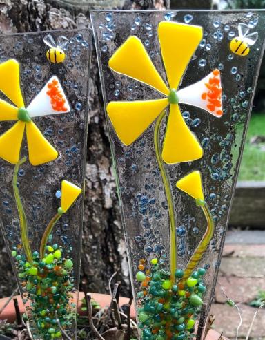 Two fused glass ornaments depicting yellow flowers sit in a flower pot outside. Partially obscured.