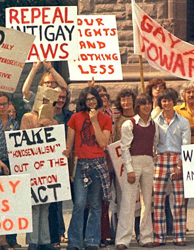A large group of people dressed in T-shirts and seventies-style flared pants, stand together holding signs with messages such as “Out of the Closet,” “Gay is Good” and “Repeal Anti-Gay Laws.” Partially obscured.