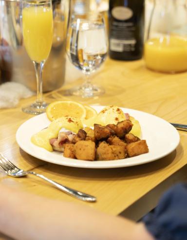 A person is sitting at a table with a glass of orange juice and a plate of eggs benedict and hash browns. Partially obscured.