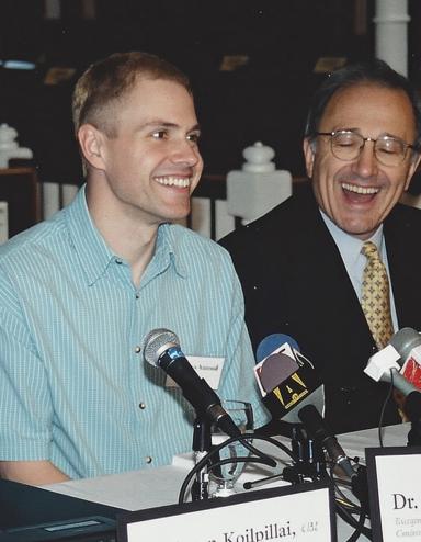 A man wearing a light blue shirt sits at a long table in front of a cluster of microphones, smiling. Seated on both sides of him are other people dressed in suits and facing microphones, also smiling. The name placards along the front of the table read, from left to right, “Murray Billet”, “Robinson Koilpillai”, and “Dr George Rodgers”, with too-blurry-to-read affiliations below the names. Another placard, knocked on its side, reads “Delwin Vriend” without an affiliation. Partially obscured.