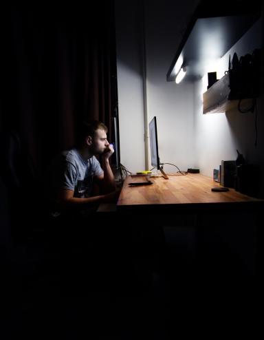 A white man with short hair sits alone at a brightly lit desk in a dark room, staring at a computer monitor. Partially obscured.