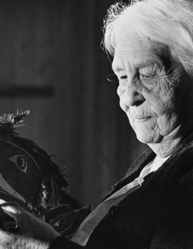 A black and white image of an elderly woman with white hair holding a mask that is shaped and painted to represent a bird’s head. It features a long beak with an open mouth and feathers attached to the top of the head. The woman is looking directly at the mask she is holding. Partially obscured.