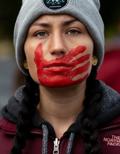 A young woman with braided hair, wearing a maroon jacket and gray winter cap, looks forward resolutely. A red handprint is painted over her mouth and face. Partially obscured.