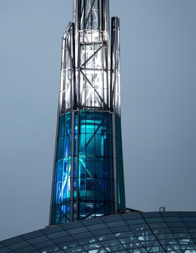 A tall, illuminated glass-and-steel spire rises above the curved patterned roof of the Canadian Museum for Human Rights, glowing in blue and white light against a soft grey sky. Partially obscured.