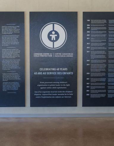 Large dark blue panels with white text and graphics displayed in the Community Corridor at the Canadian Museum for Human Rights. The centre panel features a circular logo and commemorates the 40th anniversary of the Canadian Centre for Child Protection. The two panels on either side present bilingual text in English and French. Partially obscured.