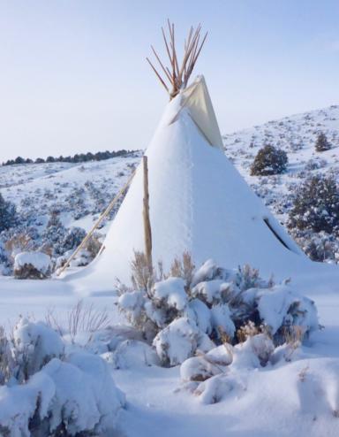 A snow-covered tipi sits in a hilly landscape dotted with trees and bushes against a blue sky. Partially obscured.