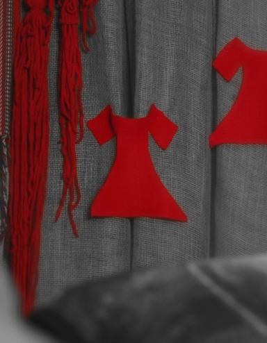 Two bright red felt dresses are pinned to a grey background next to braided red yarn and a traditional Métis sash. Partially obscured.