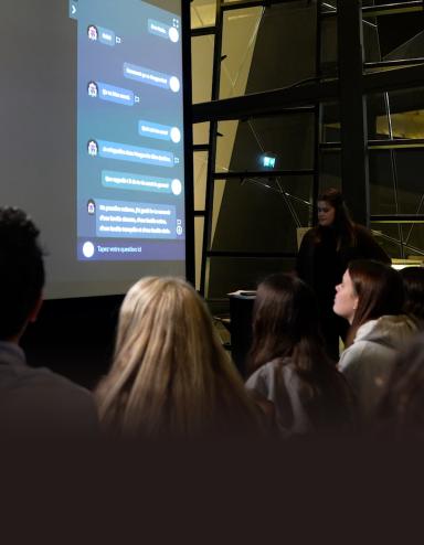 Audience members view a large projection screen displaying an elderly woman in a purple top and blue pants sitting in a white armchair, alongside an interactive message interface. The presentation takes place in a modern space with geometric glass architecture, and a presenter stands to the right of the screen. Partially obscured.