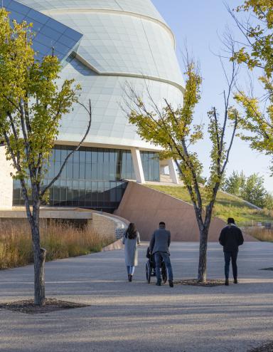 Four individuals follow the path toward the Canadian Museum for Human Rights on a fall day. Three are on foot and one is in a wheelchair. Partially obscured.