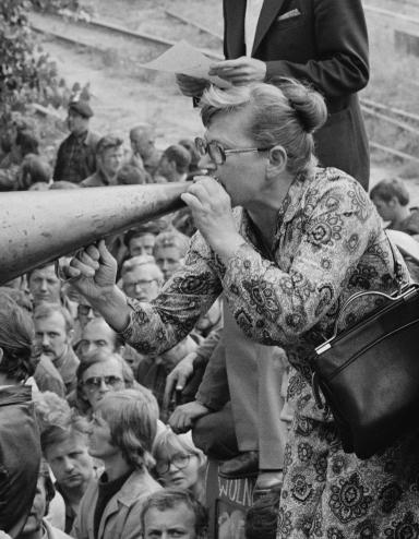 A woman wearing a dress cups her hand to a megaphone placed at her mouth. She is standing elevated above a crowd of people at a gate, with two flags waving nearby. Partially obscured.