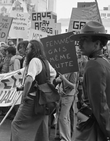A large group of protestors march through a city while two uniformed RCMP officers keep watch. In this black-and-white image, the protestors' signs and banners call for an end to discriminatory practices against members of the 2SLGBTQI+ community. Partially obscured.