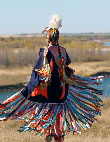 A Indigenous dancer in brightly coloured Fancy Shawl regalia beside a river. Partially obscured.