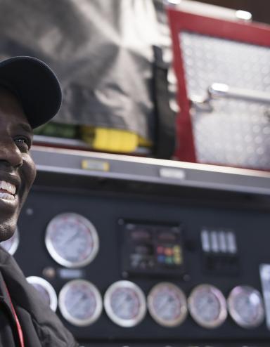 A smiling man stands in front of a fire truck. Partially obscured.